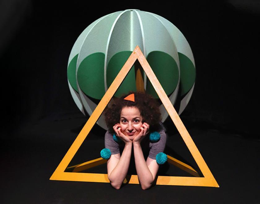 Photo of a performer crouched on the floor with their face held between their hands. They are lying through a light orange pyramid-shaped frame, with a green sphere above their head.