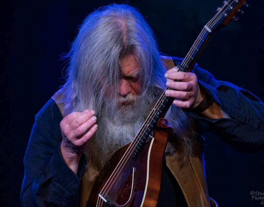 Grey haired and bearded man intently focuses on playing his mandolin against a dark blue background