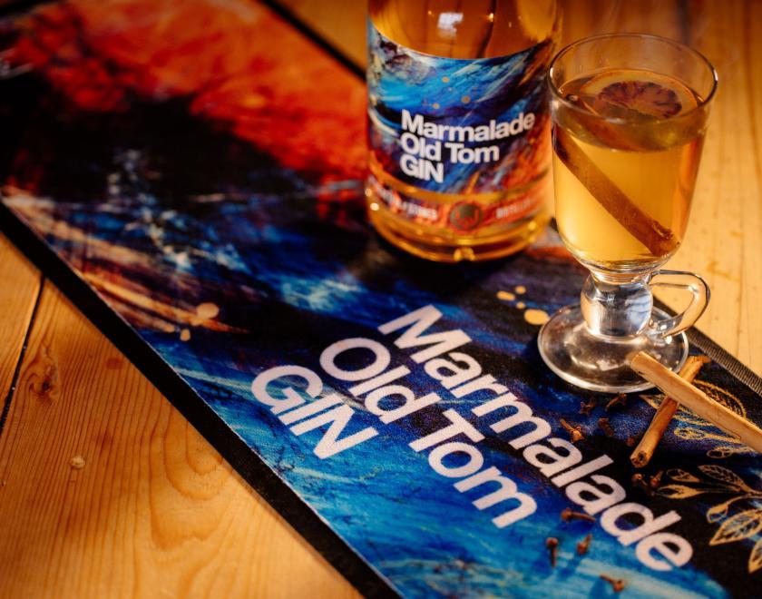 A mainly blue bar runner with the words "Marmalade Old Tom Gin" on it lies diagonally across the wooden background. On it is a bottle and a glass of liquid with a cinnamon stick in it