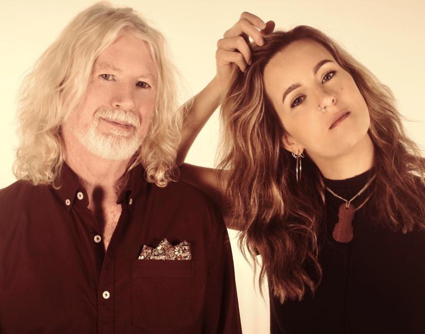 Promotional photograph of the duo We Mavericks. The duo are facing the camera and wearing black. They are standing in a casual pose. There is a light gold tinge to the photograph.