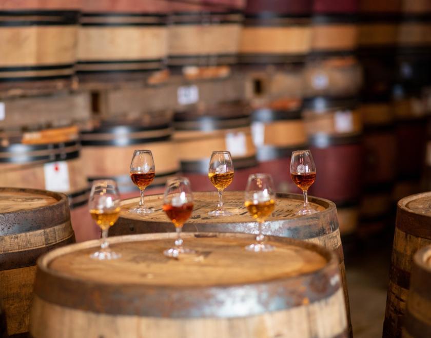 Six nosing glasses with arranged in two rows of three one behind the other on the flat tops of two large wooden casks. They are filled with different coloured rums, the row behind is in focus and the one in front is slightly blurry. The background of the picture is rows of more wooden casks