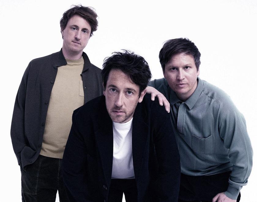 Band The Wombats, three white men in their late thirties wearing light jackets and t-shirts, looking puzzled at the camera