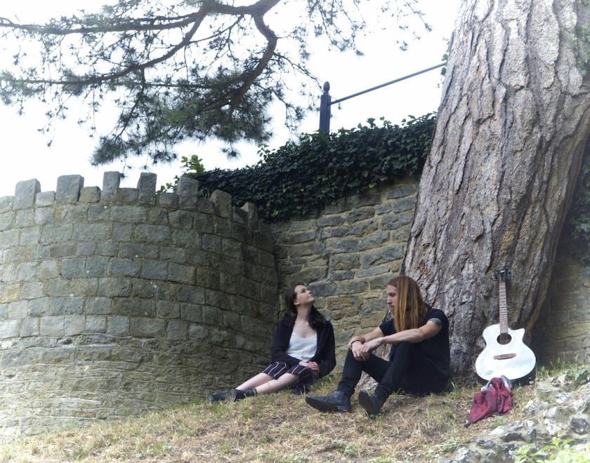 Two musicians sit with an old stone wall behind them, a guitar is propped against the trunk of a pine tree