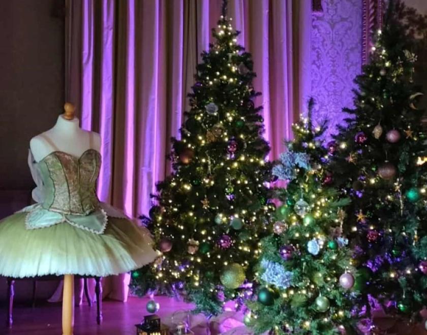 A green and pink ballet tutu sits next to three different sized christmas trees decorated in green and purple baubles. Behind the costume and trees is a purple light casting light and shadow over the group and the curtain behind. 