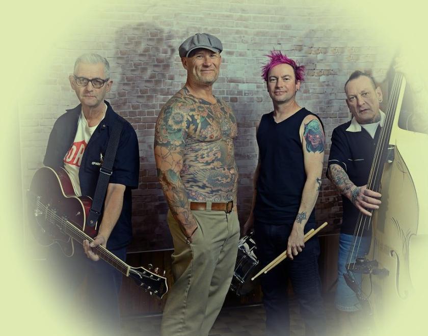 Photo of the band members of Guana Batz, four middle-aged men. Two are holding instruments, one is shirtless and covered in tattoos.