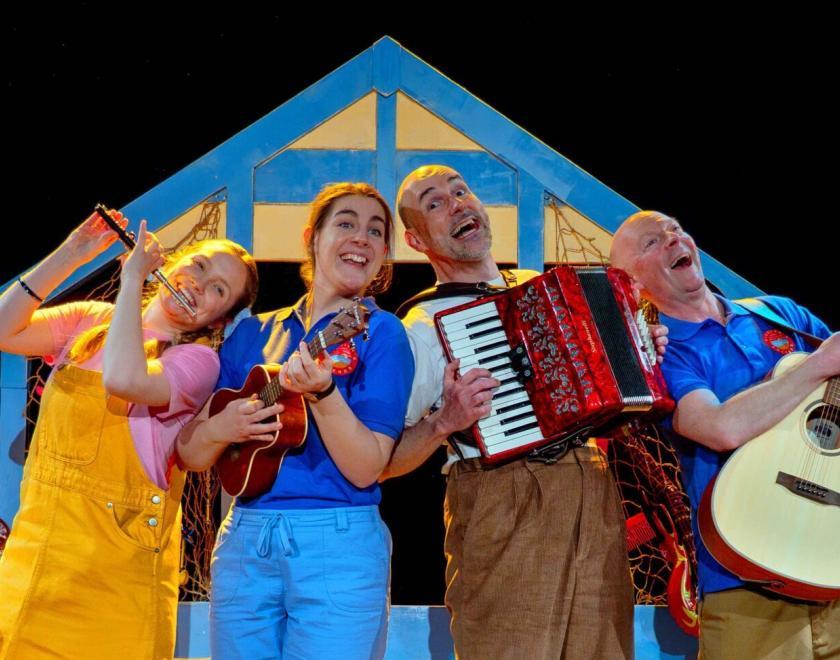 Four actor-musicians on stage wearing beach-style costumes in front of a beach hut