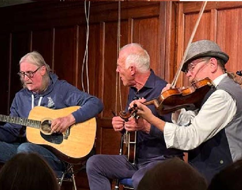 Photo of the trio Baum, Maggs and Taylor playing music in a folk club environment. They are playing guitars and fiddle.