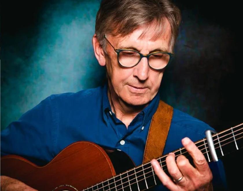 Photo of Bob Wood playing guitar. There is a spotlight on Bob's face and hands. The background is lit in blue and Bob is wearing a blue shirt.