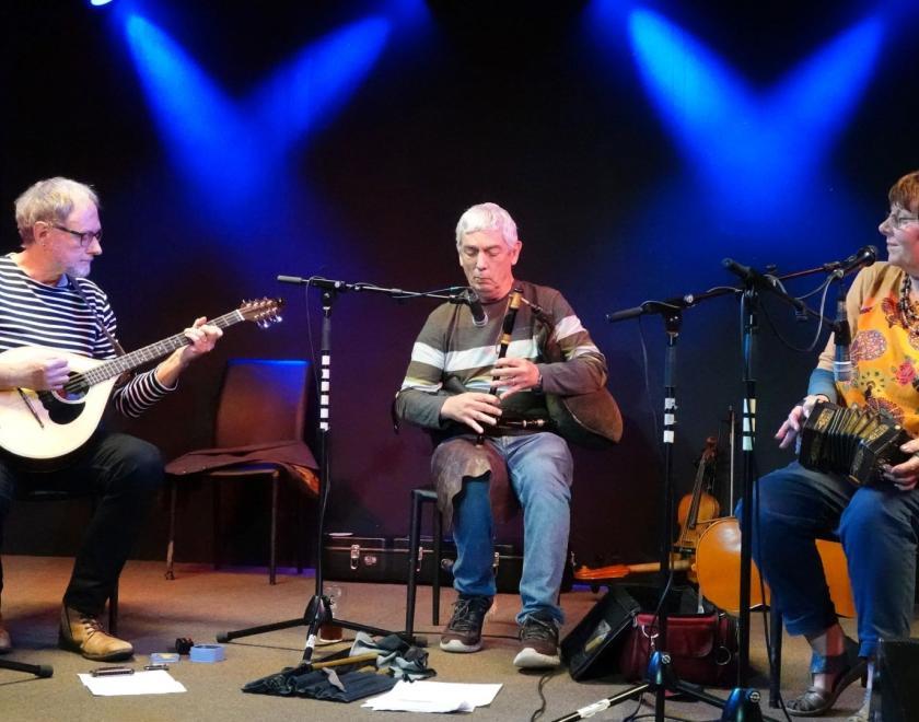 Three musicians sitting on stage, playing a bouzouki, Uillean pipes and concertina respectively