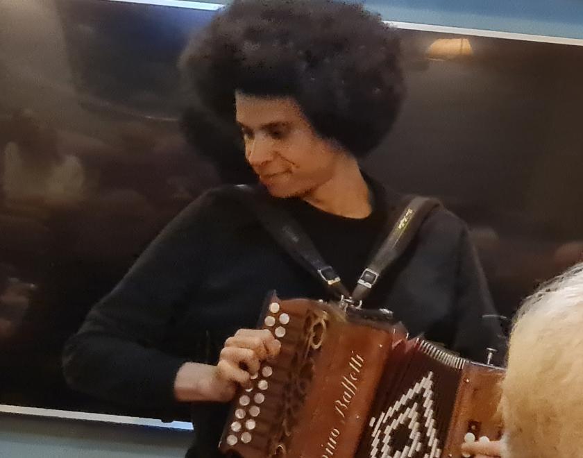 Photo shows Cohen Braithwaithe Kilcoyne playing at a folk club. Cohen is playing a melodeon. He is looking down to the side and smiling.