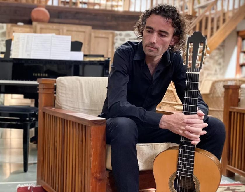 Guitarist Gerard Cousins sat holding his acoustic guitar in a library-like room with a piano in the background