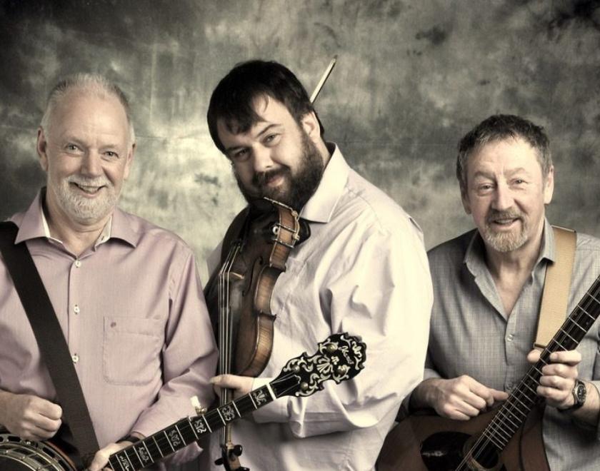 Sepia-tinted image of the three members of North Sea Gas posing in the spotlight with musical instruments (guitar, fiddle and bouzouki)