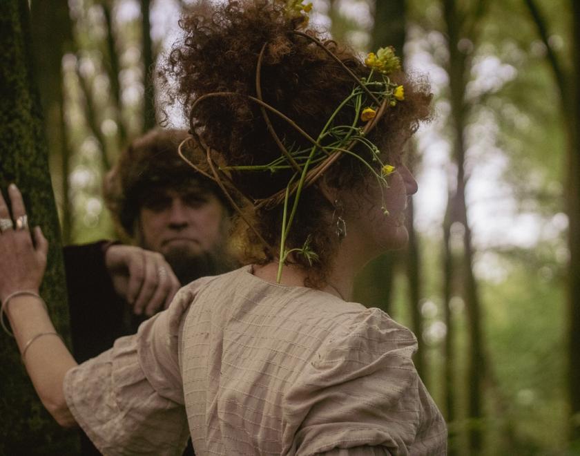 A woman with curly hair with her back to us, and a man with a big beard facing us, standing in a forest