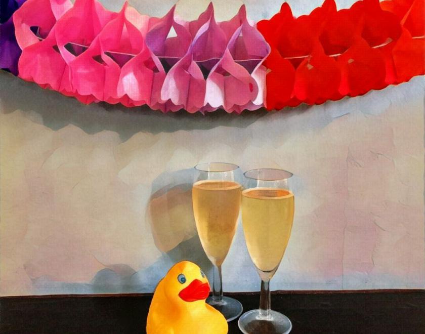 A rubber duck poses with 2 glasses of champagne against a lilac background with a paper concertina decoration above