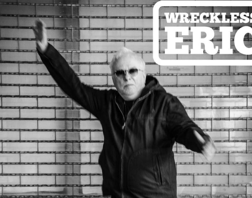 Black and white photo of Wreckless Eric, wearing all black and raising one arm in the air