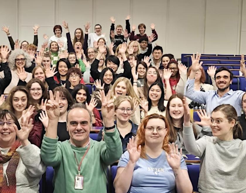 Auditorium filled with people of various ages all waving their hands at the camera
