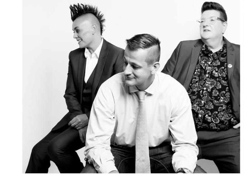 Black & White image of three seated white-skinned musicians looking to the left side. Person sat in front is wearing a white shirt and pale coloured tie, he has short hair and an amused expression; the person behind and to the left is wearing a dark satin effect suit and waistcoat and has dark spiked hair; the person behind and to the right is wearing a mid gray suit jacket and trousers with dark paisley shirt and glasses