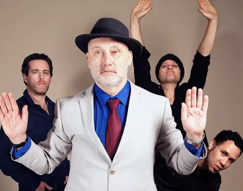 Band members standing with their hands up. Singer Jah Wobble is at the front in a white suit jacket and trilby hat