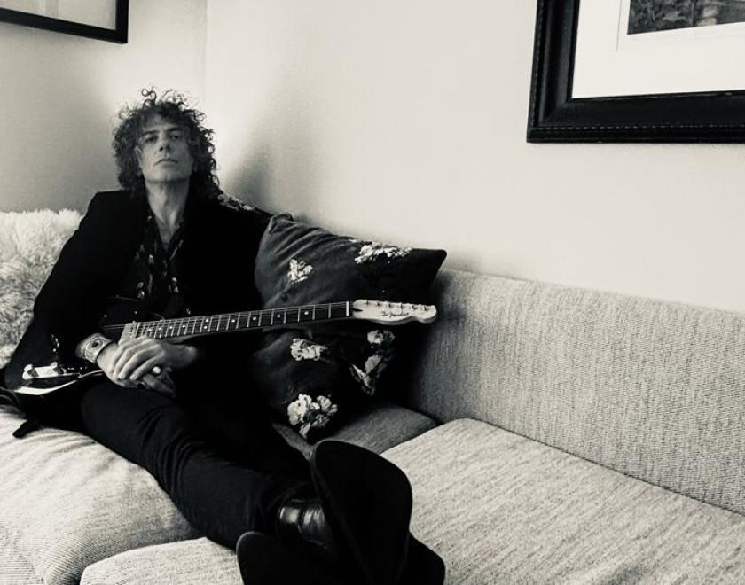 Kavus Torabi in a black suit, lounging on a white sofa with a guitar