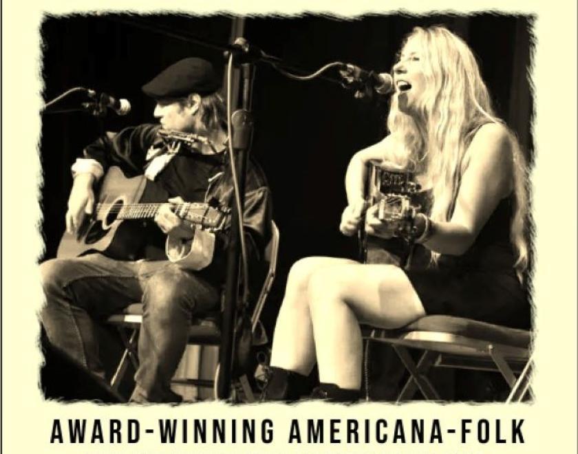 Sepia tinted black and white photo of two seated musicians. The one on the left wears jeans and a cap and is playing a guitar the one to the right has long blonde hair she is also playing the guitar and singing