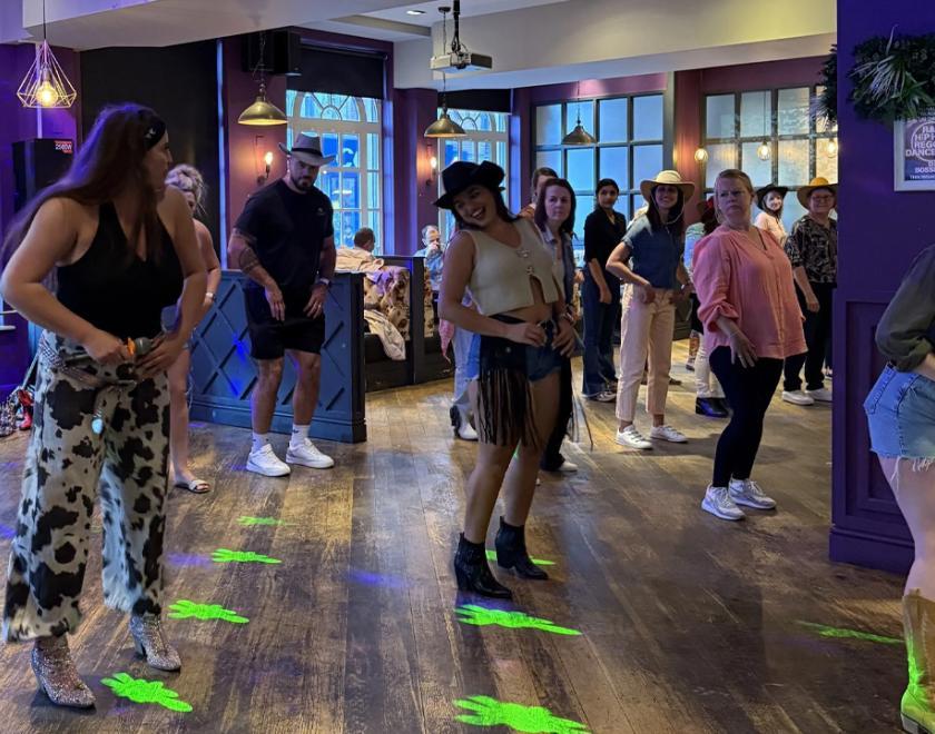 Group of people learning line-dancing steps in a bar. They are mostly wearing cowboy hats.