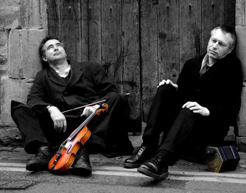 Black and white photograph of Mat Green and Andy Turner sitting in a nonchalant pose on the ground in front of an old wooden door and over double-yellow street lines. Mat has a fiddle, the only coloured object in the photo, and Andy has a concertina.