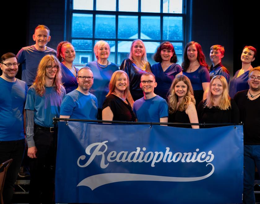 Members of the Readiophonics a capella group, all wearing blue and black outfits, standing behind a banner with their logo on it.