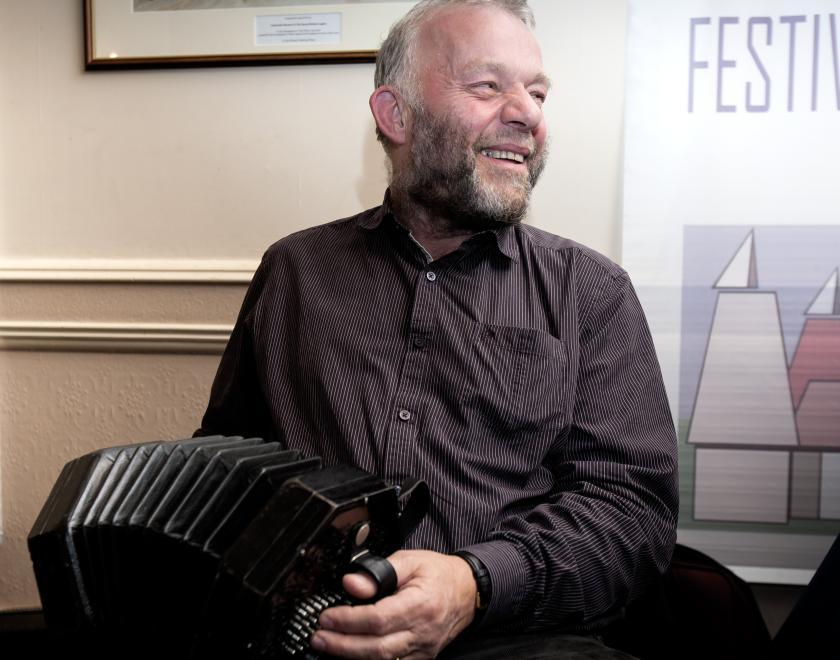 Photo of Steve Turner playing his concertina. Steve is wearing dark clothing and is sitting in a traditionally-styled room (possibly a pub) with a picture behind him and a dado rail on the wall. Steve is smiling. Steve's concertina is a duet.